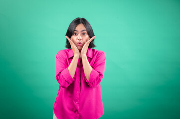 An Asian woman in a vibrant pink shirt stands against a green background, expressing surprise with her hands on her cheeks and an open-mouthed smile, capturing a lively and joyful reaction