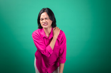 Fototapeta premium An Asian woman wearing a bright pink shirt against a green background is holding her shoulder with one hand, expressing pain or discomfort, with her face showing a pained expression