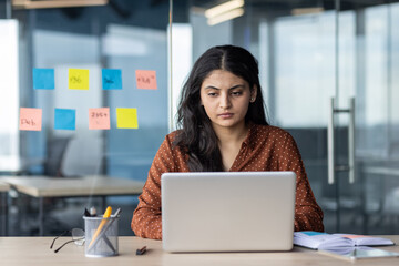 Young hispanic business woman works intently on laptop in modern office. Surrounded by colorful notes, stationery items, reflecting focus, professionalism, concentration, productivity. © Liubomir