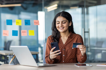 Young hispanic business woman using smartphone and credit card at office desk, analyzing business data on laptop. Concept of multitasking, financial management, technology in modern workspace.