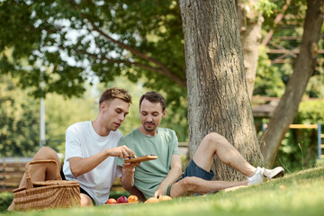 Two joyful men picnic on a sunny day, exploring treats while surrounded by nature.