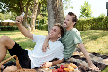 Two men enjoy a delightful picnic, smiling while taking a selfie together in a serene park.