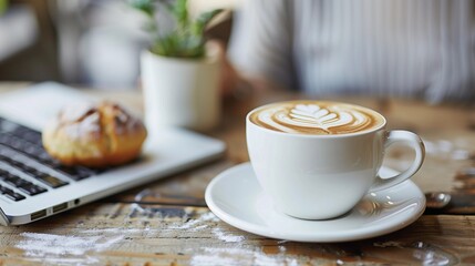 a creamy latte in a white cup next to a laptop and fresh bread on a marble surface, ideal for coffee shop, workspace, or remote work themes