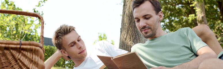 Two men sit closely in a sunny park, engrossed in a book, sharing smiles and laughter.