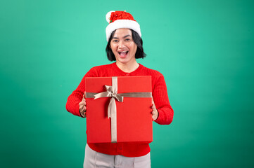 An Asian woman in a Santa hat and red sweater is smiling joyfully while holding a large red gift box with a golden ribbon against a green background, spreading holiday cheer