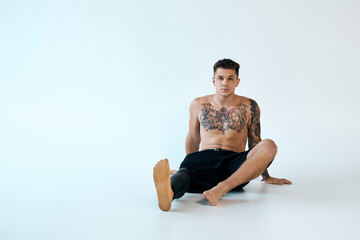 A confident young man with tattoos sits on the floor, showcasing his prosthetic leg in a well-lit...