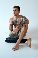 A young man with tattoos poses in a studio, highlighting his prosthetic leg and muscled form while...