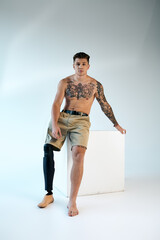 A young man with striking tattoos showcases his prosthetic leg while posing in a minimalist studio.