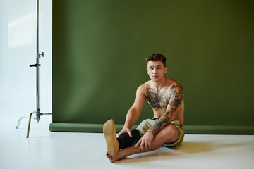 In a vibrant studio, a tattooed young man sits thoughtfully, highlighting his prosthetic leg with...