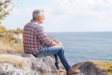 Fototapeta premium A 65 year old European grandfather sits peacefully on a cliff, gazing at the vast ocean horizon. The soft dawn light enhances the tranquil atmosphere, creating a serene moment of contemplation and con