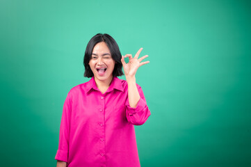 Fototapeta premium An Asian woman in a bright pink shirt poses against a green background, smiling and winking while making an 