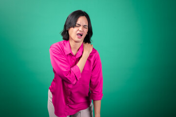Fototapeta premium An Asian woman wearing a bright pink shirt against a green background is holding her shoulder with one hand, expressing pain or discomfort, with her face showing a pained expression