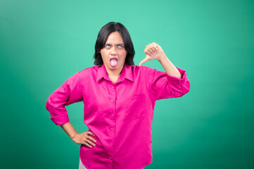 An Asian woman in a bright pink shirt poses against a green background, making a playful face by sticking out her tongue and giving a thumbs-down gesture. Her expression is humorous