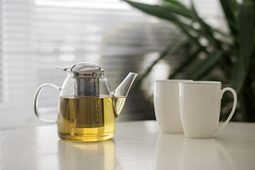 Glass teapot with brewed tea beside two white cups in bright indoor setting