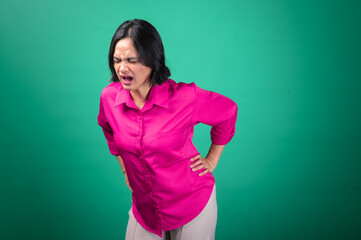 An Asian woman in a bright pink shirt against a green background expresses pain, with her hand on her back as if experiencing backache or discomfort. Her face shows an intense expression of distress.