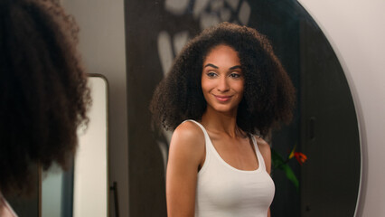 Young beautiful African American woman girl enter to bathroom looking pampering mirror reflection check hair after haircare shampoo mask balm touch curls hairdo curly enjoy hairstyle walking away