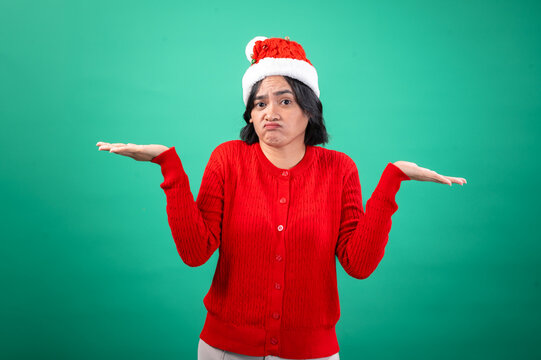 An Asian woman in a red sweater and Santa hat shrugs with both hands raised, expressing confusion or indifference, standing against a green background. Her facial expression is questionin