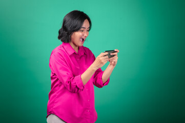 An Asian woman in a bright pink shirt looks surprised while holding her smartphone horizontally, as if playing a game or watching something on it. Her expression is focused and captivated.