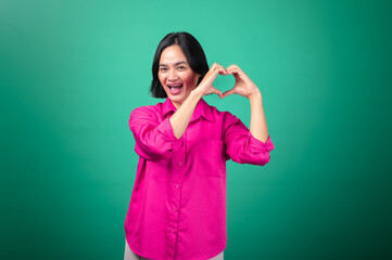 Fototapeta premium An Asian woman in a pink shirt stands against a green background, forming a heart shape with her hands in front of her chest and smiling warmly, conveying love, positivity, and affection.