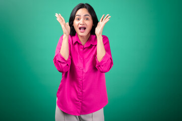 Fototapeta premium An Asian woman in a bright pink shirt stands against a green background, expressing surprise with an open-mouthed smile, her hand raised to her face in a playful, animated gesture