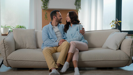 Cheerful beautiful young couple in love hugging cuddling relaxing on couch at modern home touch...