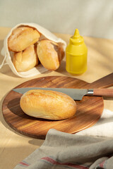 A rustic food scene featuring a freshly baked bread roll on a wooden cutting board, accompanied by a kitchen knife and a mustard bottle. Warm morning light accentuates the golden crust