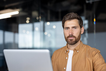 Mature businessman with a confident expression using laptop in modern office. He exudes professionalism, focus, determination, dressed in casual attire, working on business tasks.