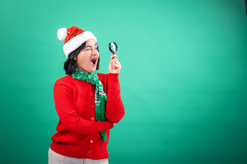 An Asian woman dressed in a Santa hat, red sweater, and green scarf, holding a magnifying glass and winking, set against a green background, showcasing a playful and curious holiday spirit
