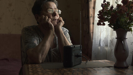Thoughtful sad elderly woman listening to portable radio while sitting by window