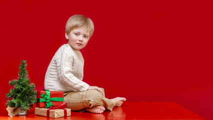 A child with blonde hair and blue eyes sits on a table next to a Christmas tree and boxes of gifts...