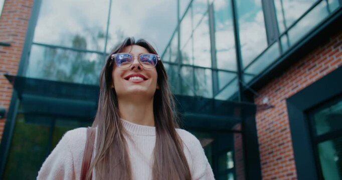 Charming Caucasian girl with glasses and straight hair walking past modern building with brick walls and tall windows. Smiling while looking straight ahead. Going to planned event for evening.