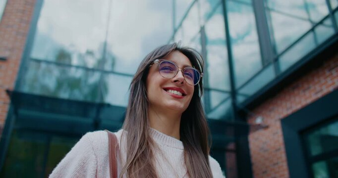 Positive Caucasian woman confidently walking away from office building. Leaving work early. Smiling while looking around. Happy about upcoming weekend. Wearing round glasses and handbag.