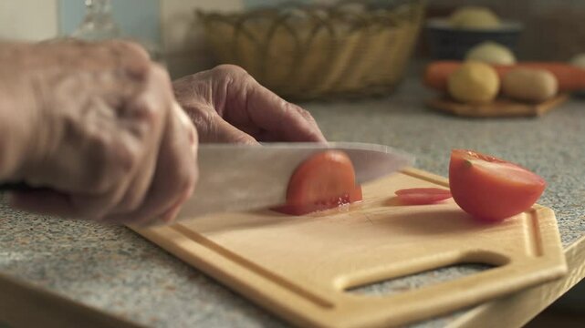 Elderly woman cutting tomato while sitting in modest kitchen. Hands close-up