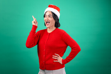 An Asian woman in a red sweater and Santa hat poses against a green background, smiling widely and pointing upwards with one hand while resting the other on her hip, adding a festive and joyful