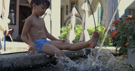 Young boy sitting at the edge of a stone fountain, splashing water with his feet, creating large splashes, enjoying playful summer fun in 800 fps slow motion