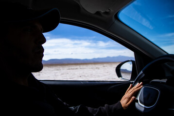 A young man's tanned hand on the black steering wheel of his car driving through Argentina's spectacular Salinas Grandes on a blue-sky day.