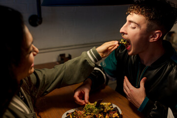 Young woman shoves food into her boyfriend's mouth while sharing a delicious plate of Mexican nachos at a restaurant during a romantic date.
