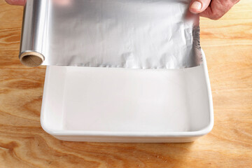 The cook spreads baking foil in a ceramic roasting pan.