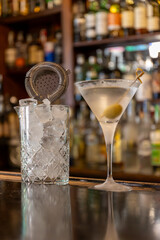 close up shot of a martini cocktail with olive in the typical martini glass, on the wooden counter of a bar with tumbler and strainer. Adult beverage, leisure, relaxation mood, italian aperitif.