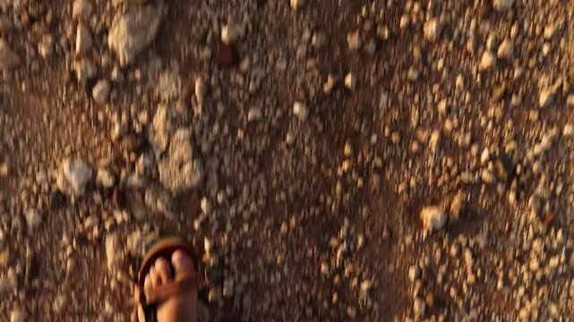 A man&rsquo;s sandaled feet trek the rocky, sandy path of Capo Sperone, Sardinia, as golden light casts warm hues on his journey.
