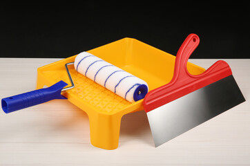 Paint roller, tray and putty knife on light wooden table against black background. Construction...