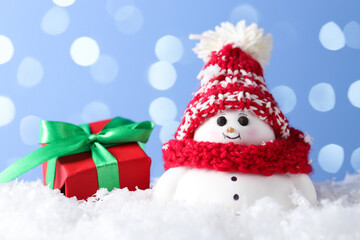Cute decorative snowman and gift box on artificial snow against light blue background with blurred lights, closeup