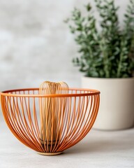 Orange wire bowl with a bamboo whisk resting on a white surface with a potted plant behind it.