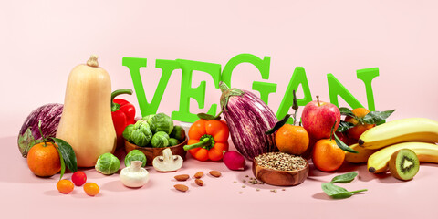 The word VEGAN in large green letters on pink table, surrounded by fresh fruits, vegetables, and a bowl of lentils