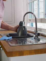 Closeup of a senior man’s hand holding a kettle under running water from a tap over a kitchen sink near a partially open sash window.
