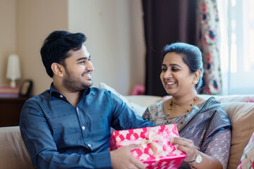 A young Indian man presenting a gift to his smiling mother in a living room, reflecting a joyful family moment.