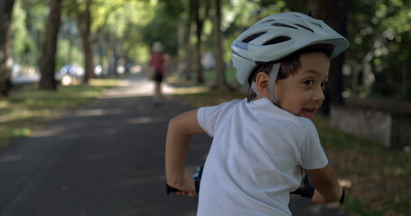 Young boy cycling with a helmet, looking back with a playful expression, on a tree-lined path...