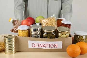 Food donation. Woman with box of different products at wooden table indoors, closeup