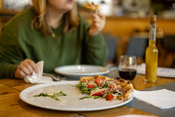 A woman holds a piece of pizza in hands. Focus on a pizza. Pizza with prosciutto, ham, arugula, tomatoes and parmesan. Italian cuisine. Food banner