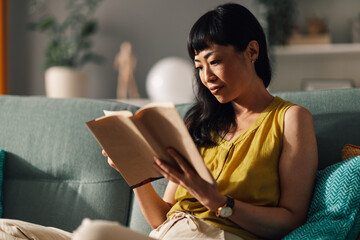 Naklejka premium Asian woman sitting on the couch reading a book in living room light.